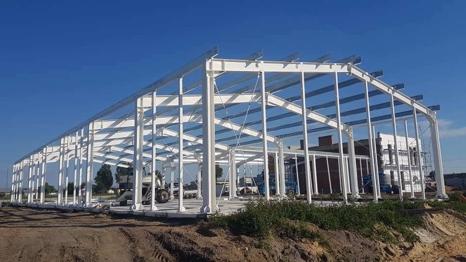Szkielet konstrukcji stalowej hali przemysłowej pomalowany na biało, widoczny na tle błękitnego nieba, z częściowo przygotowanym podłożem.