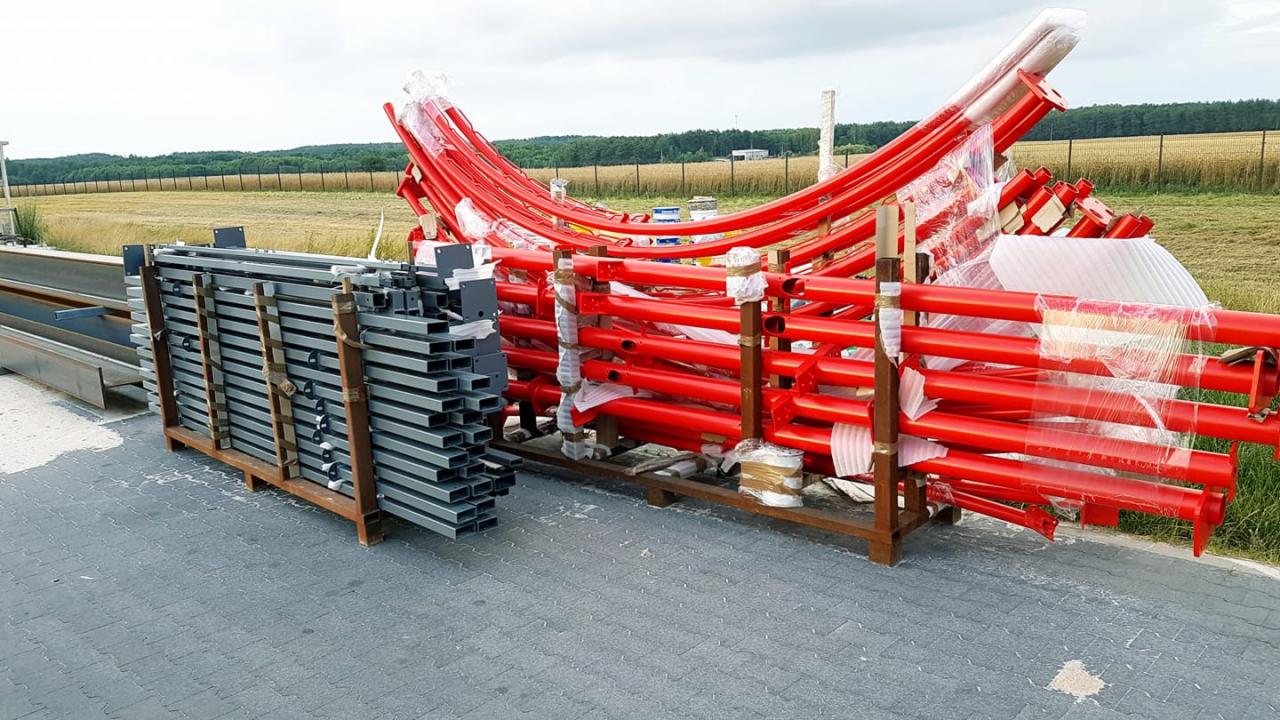 Stalowe profile w kształcie prostokąta i rury pomalowane na czerwono, zabezpieczone folią, ułożone na paletach, widoczne pole w tle.