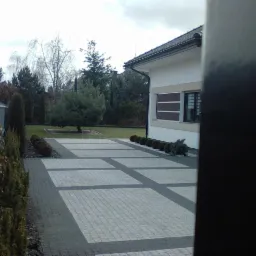 Ułożona kostka brukowa w geometryczny wzór na podjeździe, obok zadbany trawnik i krzewy, widok częściowo zasłonięty elementem ogrodzenia.