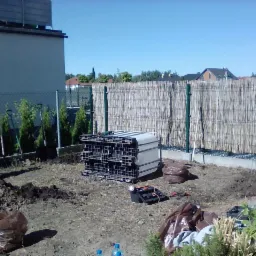 Przygotowanie terenu pod trawnik z rolki: pracownik wyrównuje ziemię, w tle rząd tuj w doniczkach i paleta z rolkami trawy, widoczna bambusowa osłona ogrodzenia.