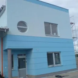 Dwukondygnacyjny budynek po dociepleniu i pomalowaniu elewacji na biało i błękitno, z widocznymi rusztowaniami i pracownikiem przy ścianie.