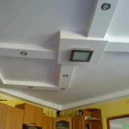 Podwieszany sufit z dekoracyjnymi elementami gipsowymi, oświetleniem punktowym i centralnym panelem z brązową ramą, widok z dołu na tle żółtej ściany i drewnianych szafek kuchennych.