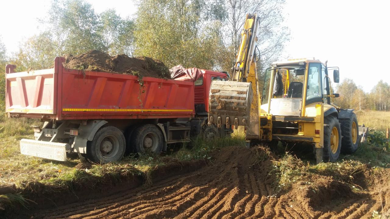 Żółta koparko-ładowarka nabiera ziemię do czerwonej ciężarówki na tle drzew i pola w słoneczny dzień. Ślady opon na ziemi.