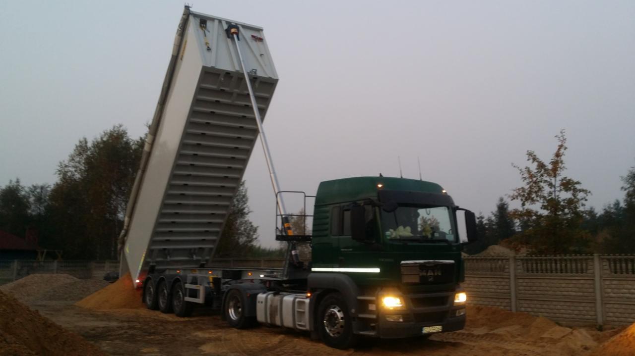 Ciężarówka MAN z otwartą naczepą wysypuje piasek w Wierzchowisku, w tle widoczny betonowy płot i drzewa.