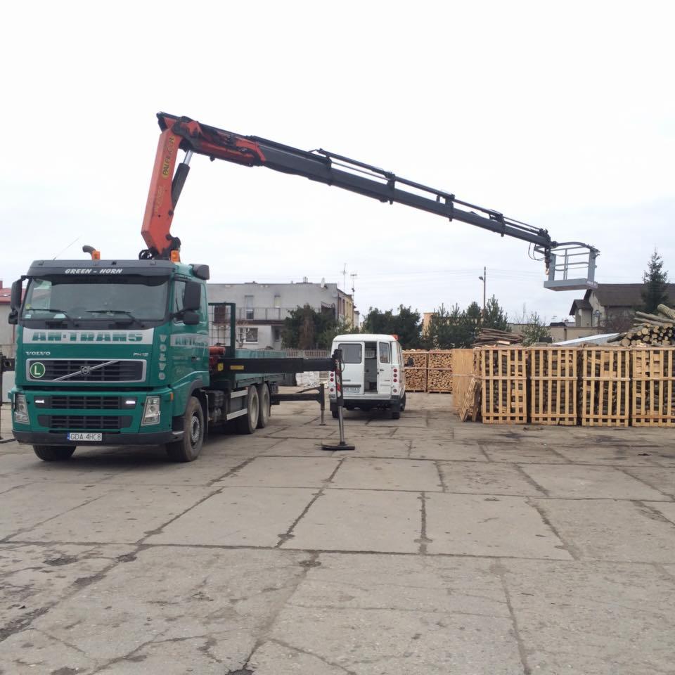 Ciężarówka z dźwigiem koszowym marki Volvo, z wysięgnikiem uniesionym nad stertą drewna opałowego, na betonowym placu.