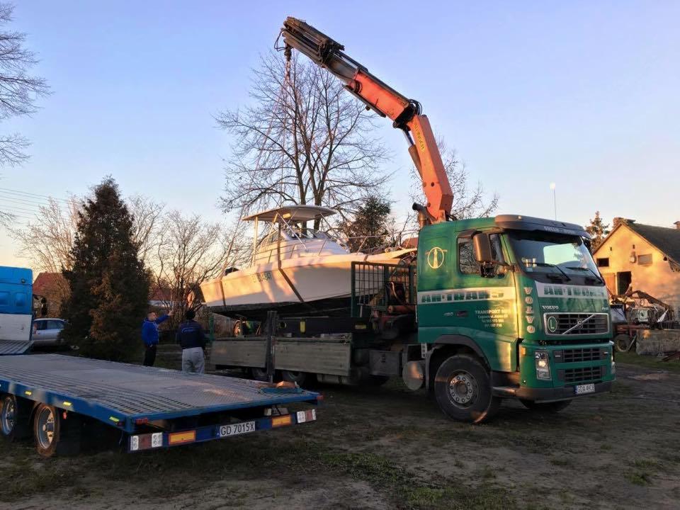 Zielony samochód ciężarowy Volvo z dźwigiem załadowuje białą łódź na przyczepę; widoczni dwaj mężczyźni obserwujący operację, w tle budynek mieszkalny i drzewa bez liści.