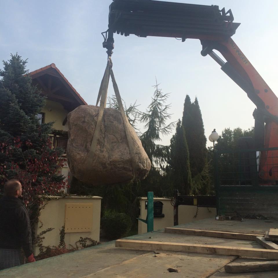 Ciężki, beżowy głaz podnoszony przez pomarańczowy dźwig Palfinger na tle domu jednorodzinnego i drzew.