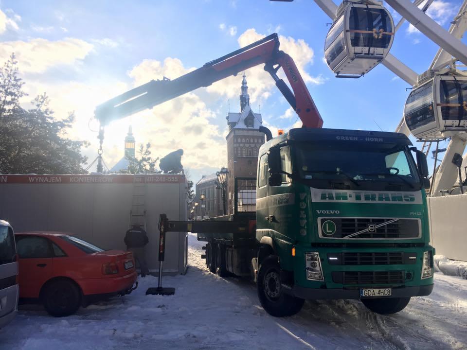 Zielona ciężarówka Volvo z dźwigiem podnosi kontener na tle zimowego krajobrazu Gdańska z kołem młyńskim i wieżą w tle.