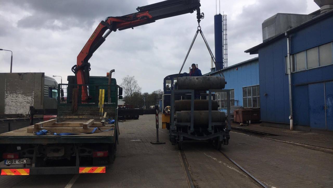 Załadunek wagonu kolejowego za pomocą dźwigu samochodowego na tle przemysłowego budynku z niebieską elewacją.