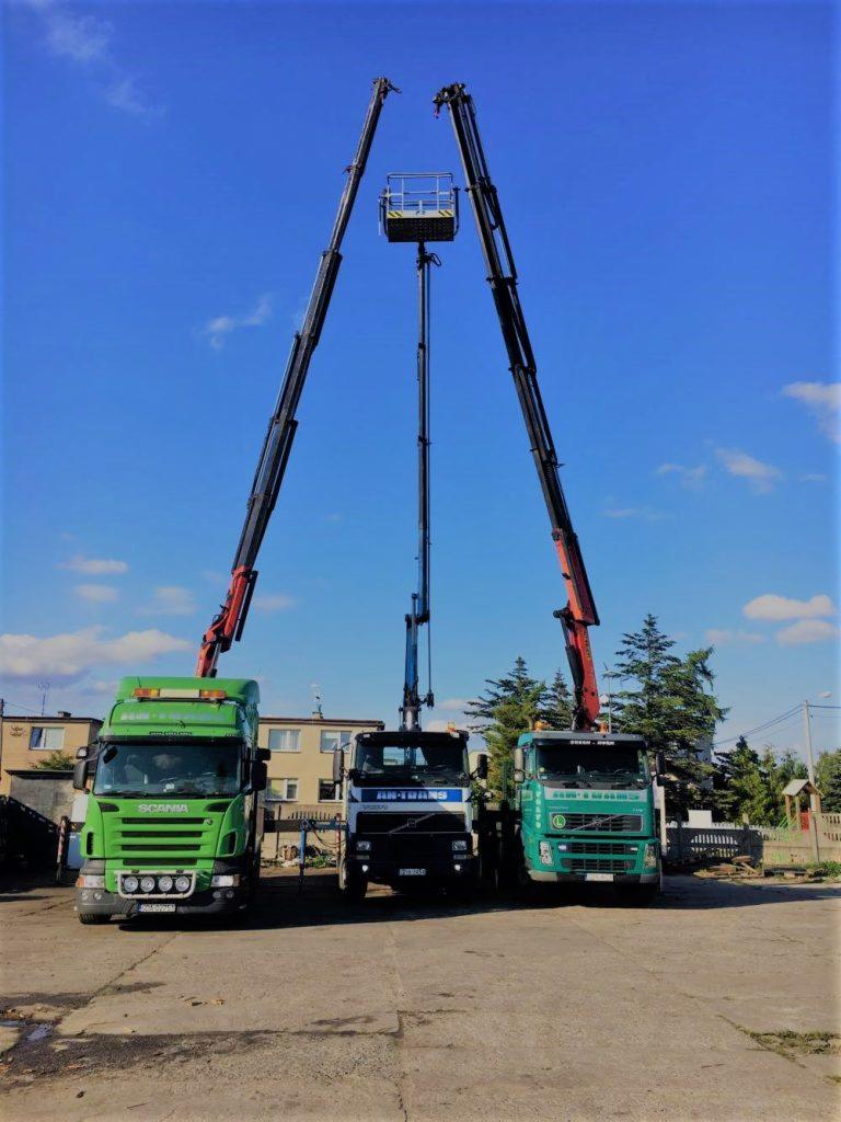 Trzy ciężarówki z podnośnikami koszowymi ustawione obok siebie na placu, dwie zielone Scanie po bokach i biała Volvo pośrodku, wszystkie z wysuniętymi ramionami podnośników w kształcie litery V.