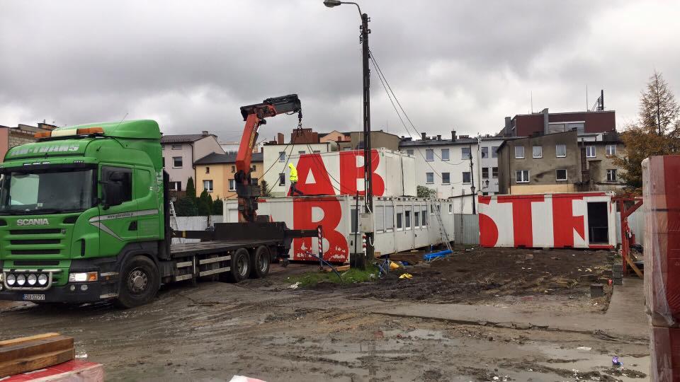 Zielony samochód ciężarowy z dźwigiem transportuje białe kontenery z czerwonymi literami na placu budowy w pochmurny dzień.