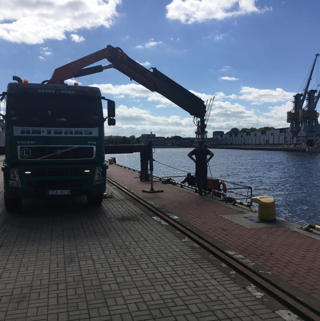 Zielony samochód ciężarowy Volvo z dźwigiem na nabrzeżu portowym, w tle widać inne dźwigi i budynki portowe, a także dwóch mężczyzn przy pracy.
