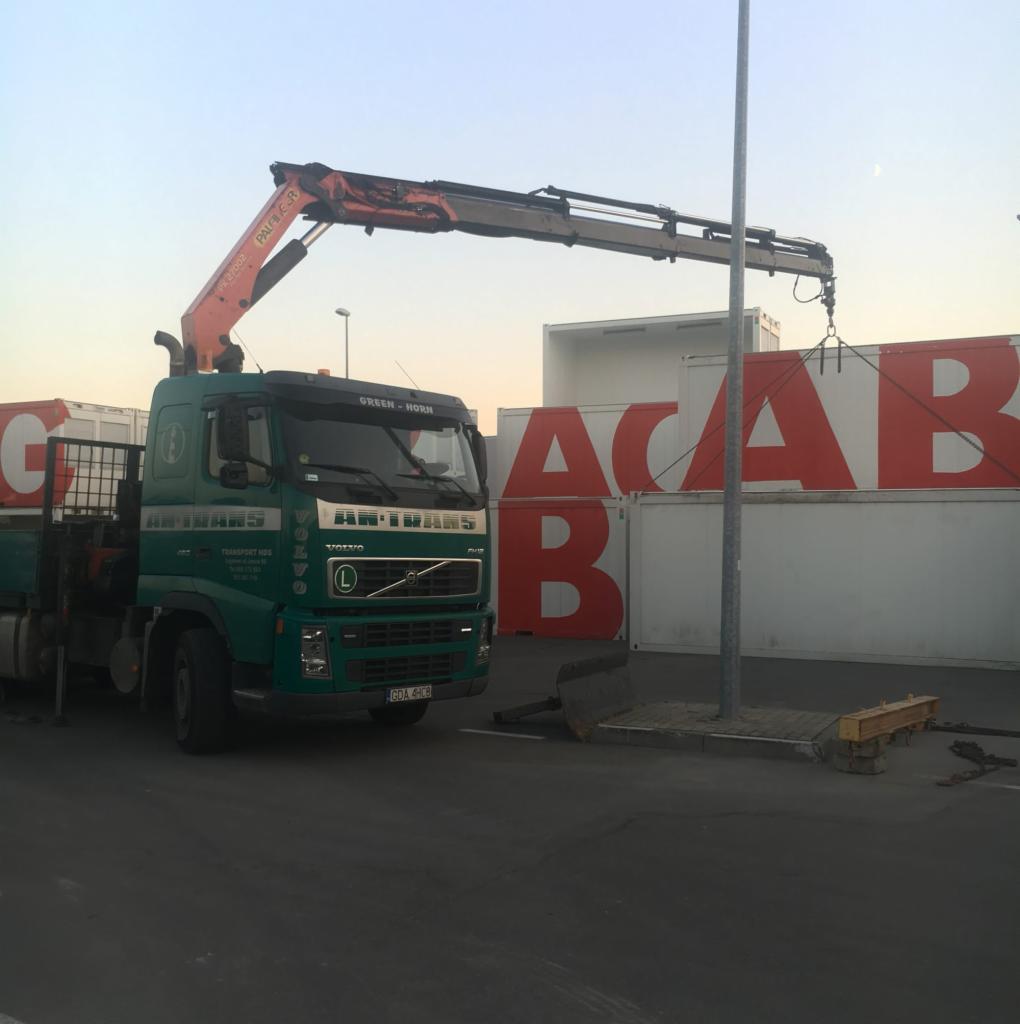 Zielona ciężarówka z dźwigiem Palfinger ładuje białe kontenery z czerwonym napisem 'ACAB' w słoneczny dzień.