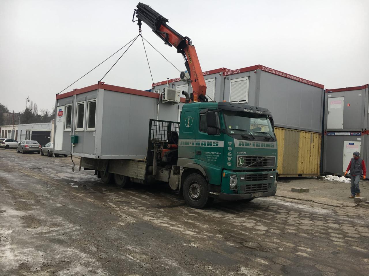 Zielona ciężarówka HDS z pomarańczowym dźwigiem podnosi kontener biurowy z klimatyzacją na tle innych kontenerów i szarego nieba.