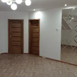 Wnętrze z trzema drewnianymi drzwiami, parkietem ułożonym w jodełkę i dekoracyjnym lustrem w romby na ścianie.