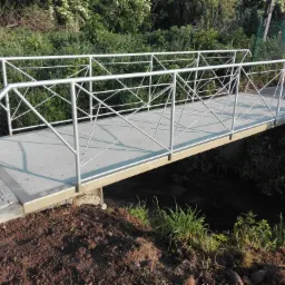 Nowo wybudowany betonowy mostek z metalowymi barierkami nad strumieniem, otoczony zielenią i świeżo ułożoną kostką brukową na podejściu.