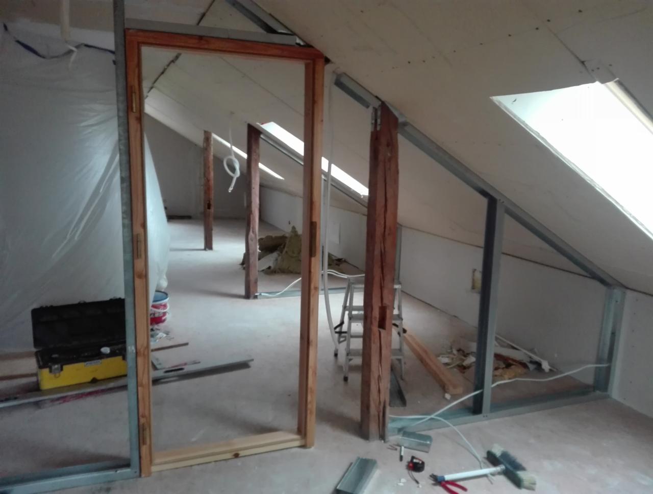 Poddasze w trakcie ocieplania i zabudowy płytami gipsowo-kartonowymi, widoczne metalowe profile, drewniana konstrukcja, narzędzia i materiały budowlane, okno dachowe.