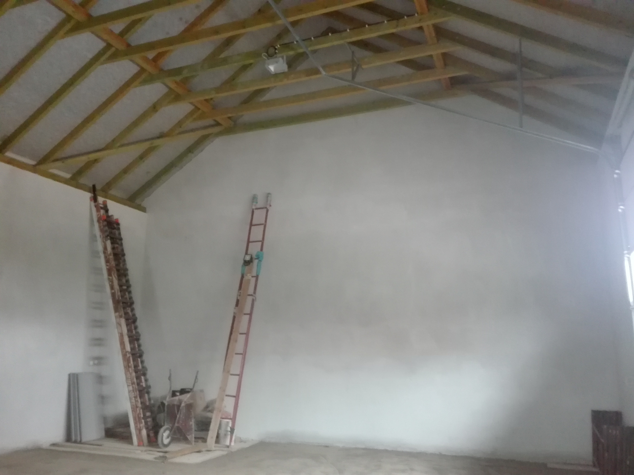 Wnętrze budynku w trakcie prac wykończeniowych: widoczne surowe ściany, drewniana konstrukcja dachu, dwie drabiny opierające się o ścianę, materiały budowlane i taczka.