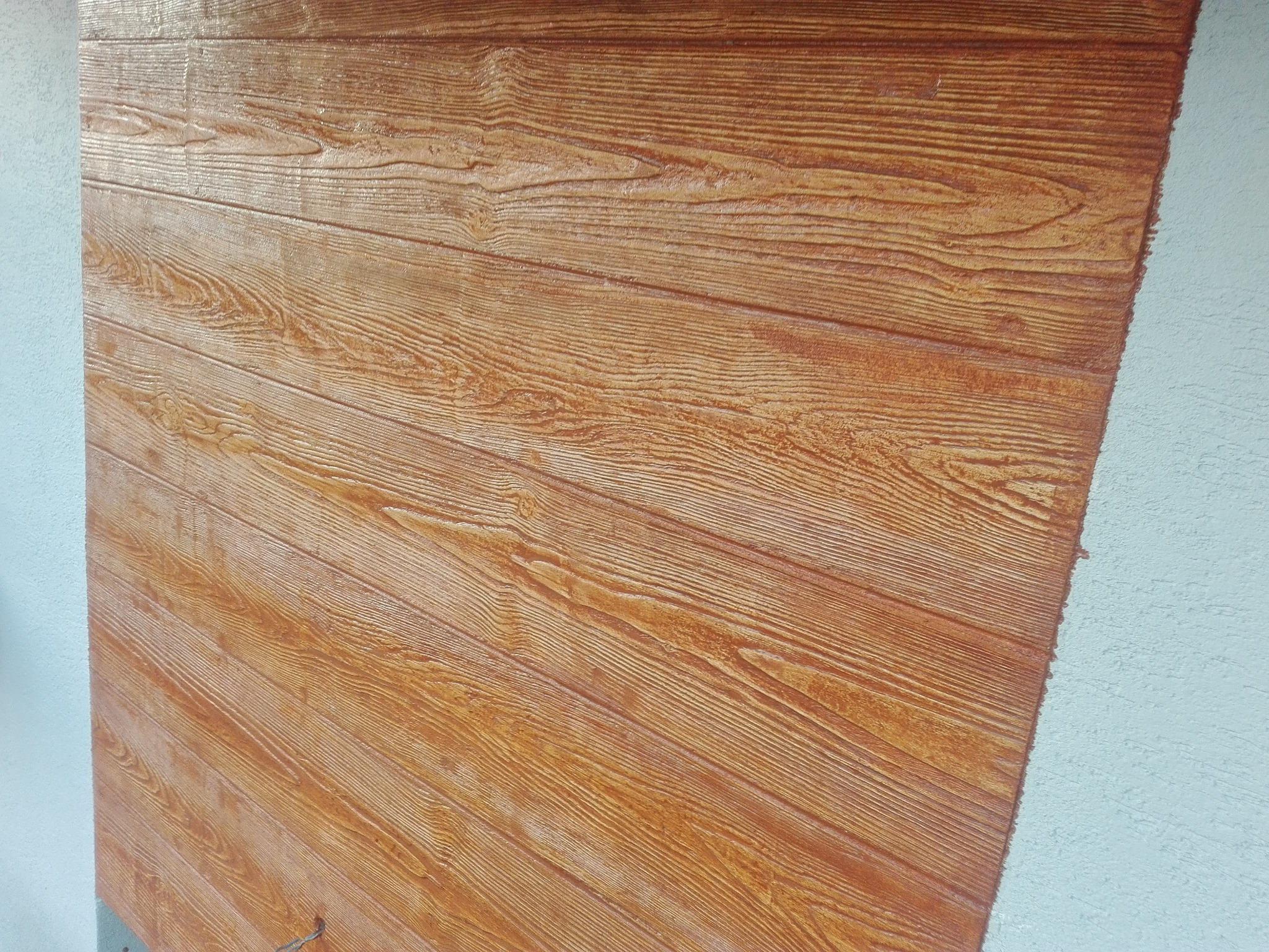 Tekstura drewna na elewacji budynku, imitacja desek ułożonych pod kątem, widoczna krawędź z jasnym tynkiem.