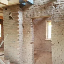 Wnętrze budynku w trakcie renowacji z odsłoniętą cegłą, widoczne drewniane elementy konstrukcyjne stropu, drabina i postać kobiety w oddali przy oknie.