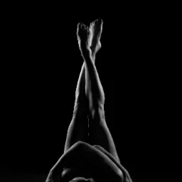 Czarno-białe, artystyczne ujęcie leżącej kobiety z uniesionymi i skrzyżowanymi nogami na ciemnym tle, podkreślające kontury ciała i grę światłocieni.