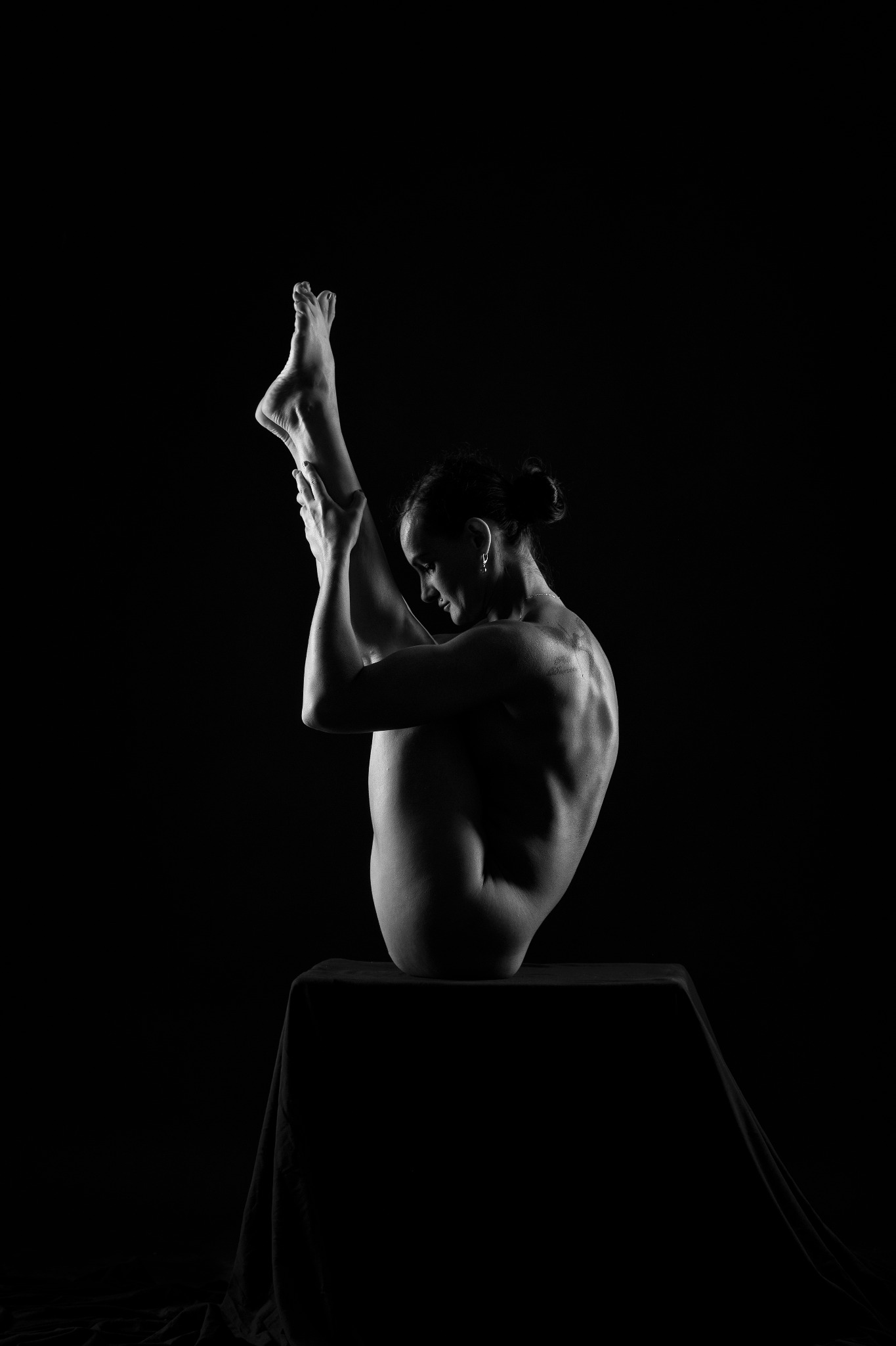 Czarno-białe zdjęcie kobiety w trudnej pozycji jogi, siedzącej na blacie obitym materiałem, z nogami uniesionymi do góry i rękami trzymającymi stopy, na ciemnym tle.