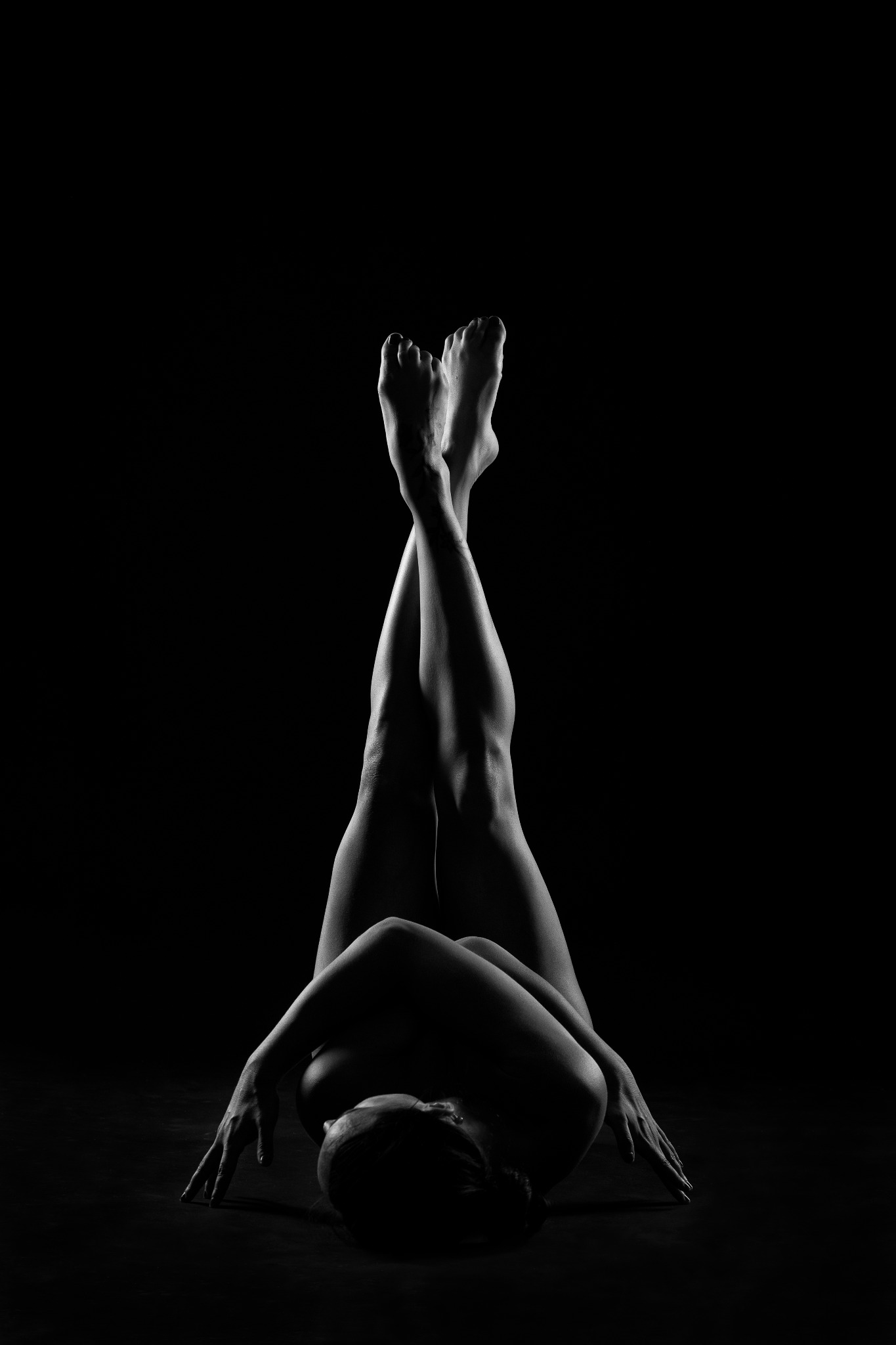 Czarno-białe, artystyczne ujęcie leżącej kobiety z uniesionymi i skrzyżowanymi nogami na ciemnym tle, podkreślające kontury ciała i grę światłocieni.