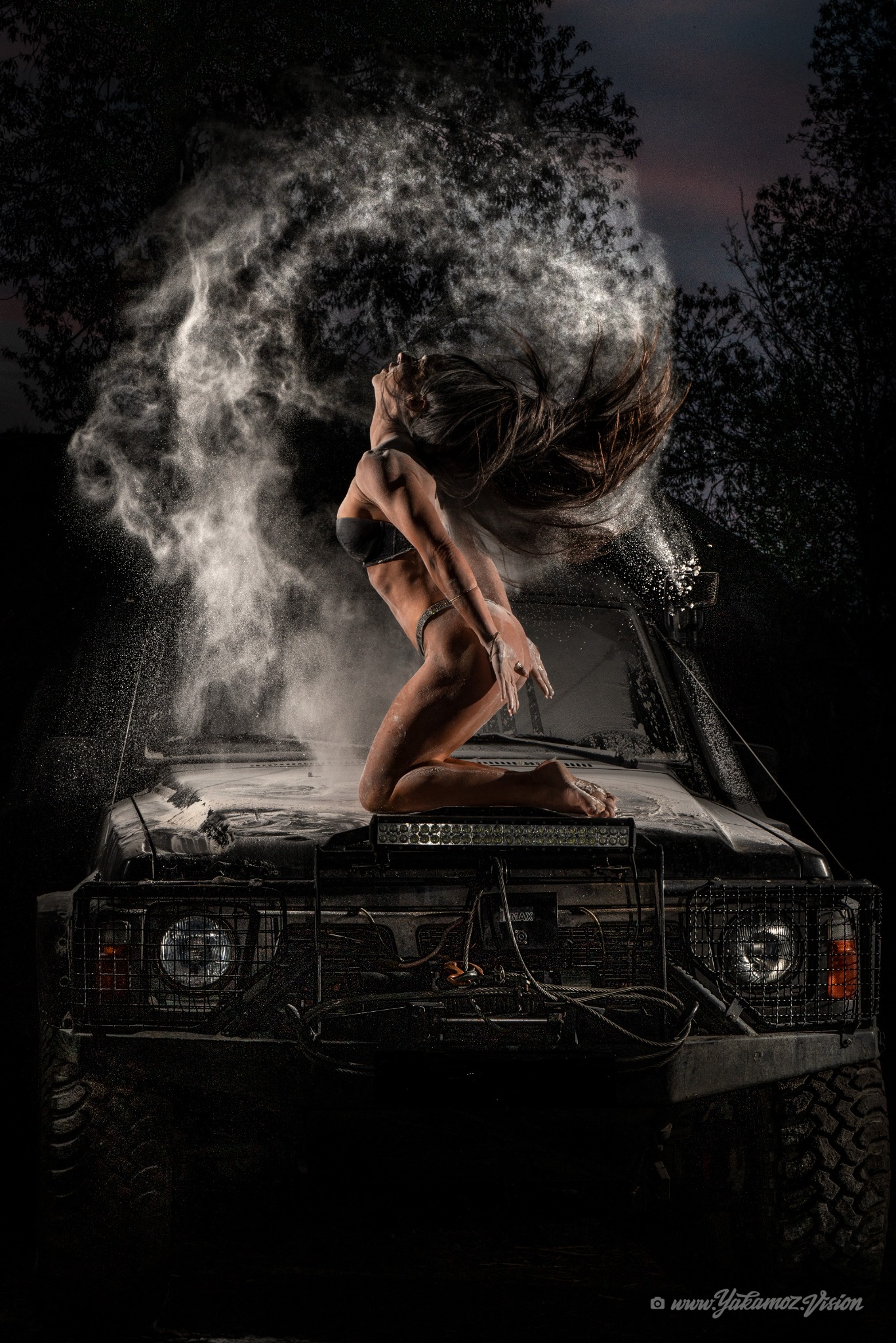 Kobieta w bikini klęczy na masce terenowego samochodu, wyrzucając włosami chmurę białego proszku w mrocznym, leśnym otoczeniu.