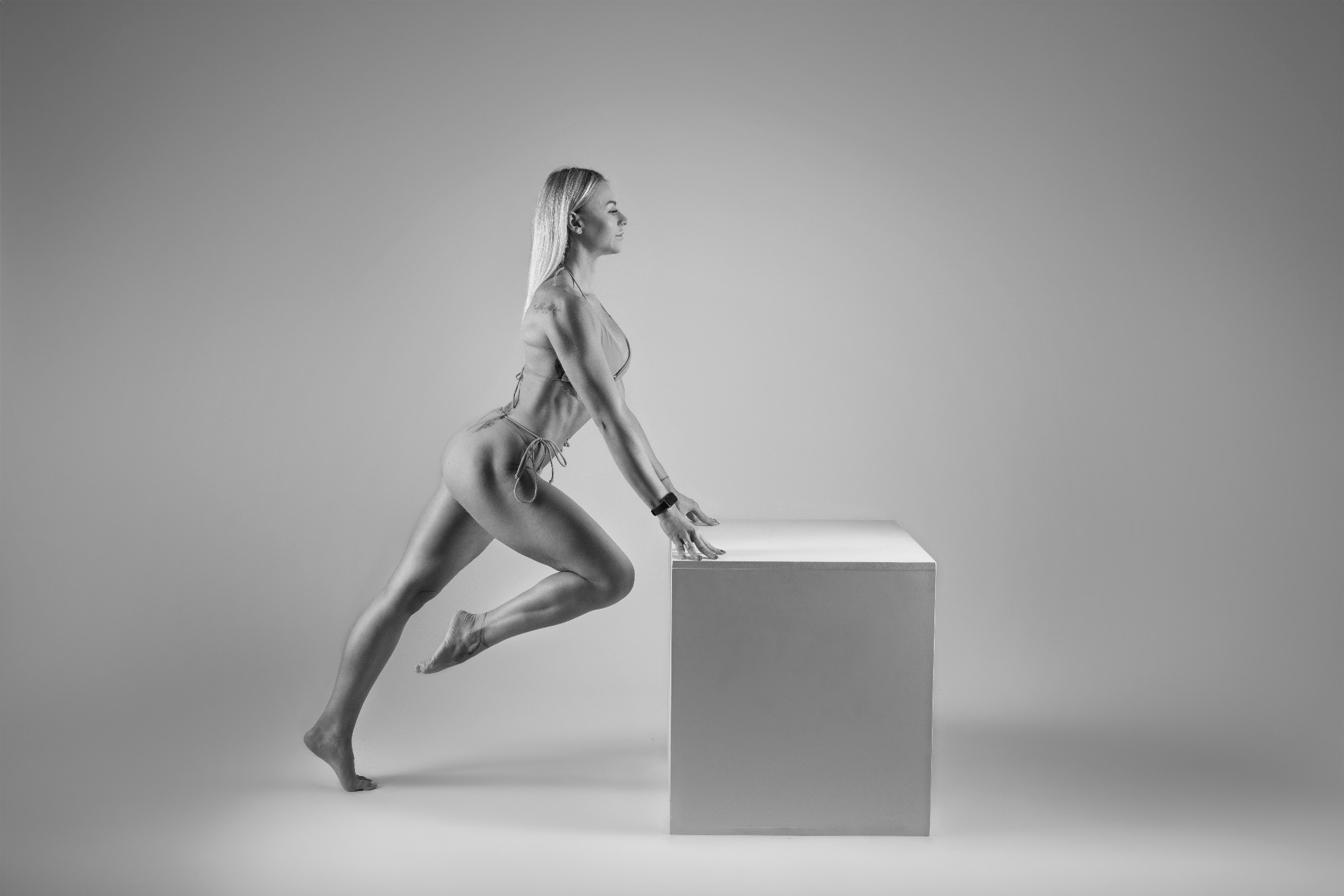 Czarno-białe zdjęcie kobiety w bikini, opierającej się dłońmi o biały sześcian, z widocznym tatuażem na ramieniu i zegarkiem na nadgarstku.