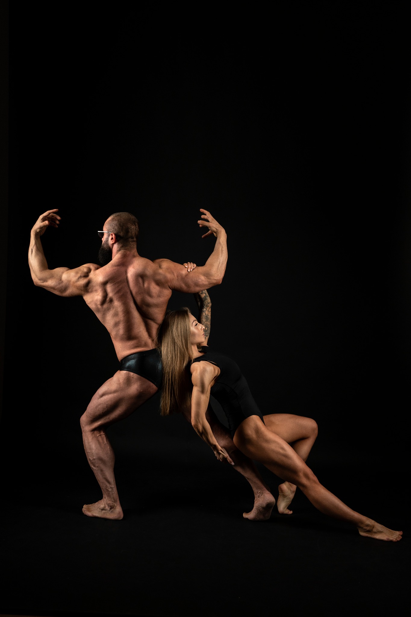 Mężczyzna z brodą i okularami oraz blondynka w czarnych strojach pozują prezentując muskulaturę na czarnym tle.