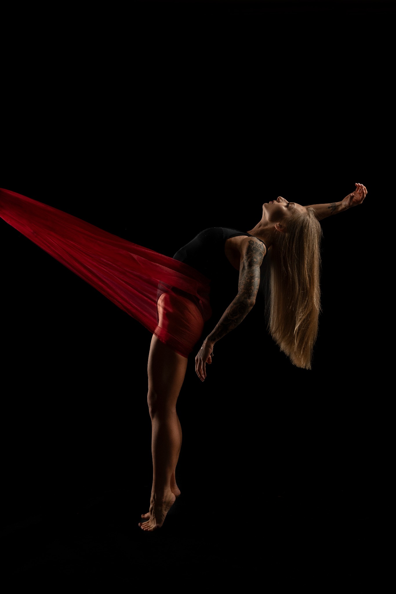 Kobieta z tatuażami, w czarnym topie i czerwonej, prześwitującej tkaninie, odchylona do tyłu na czarnym tle, z głową zadartą do góry i wyciągniętą ręką.