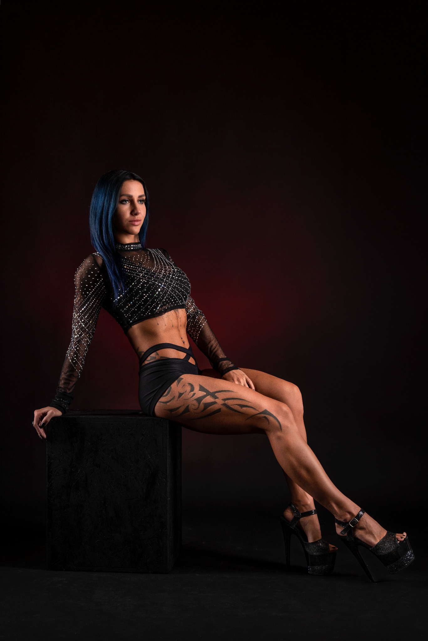 Kobieta z niebieskimi włosami w błyszczącym topie i czarnych szortach, siedząca na czarnym sześcianie, prezentująca tatuaże na udzie i wysokie platformy na tle ciemnoczerwonego studia.