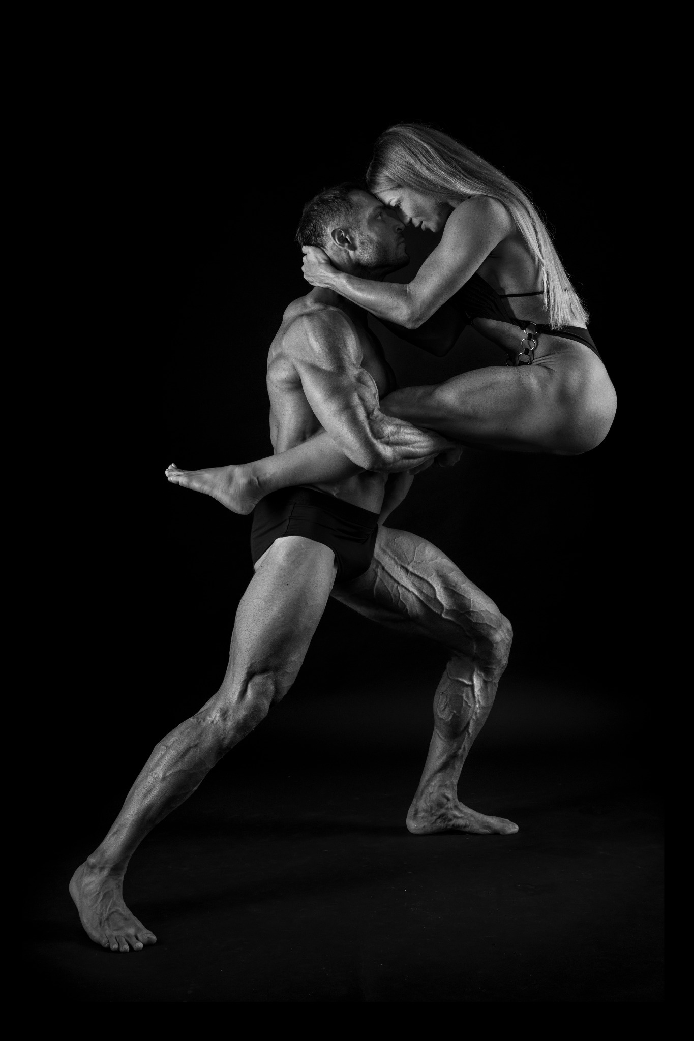 Czarno-białe zdjęcie muskularnego mężczyzny trzymającego na rękach kobietę w stroju kąpielowym na tle czarnego tła, oboje patrzą na siebie.