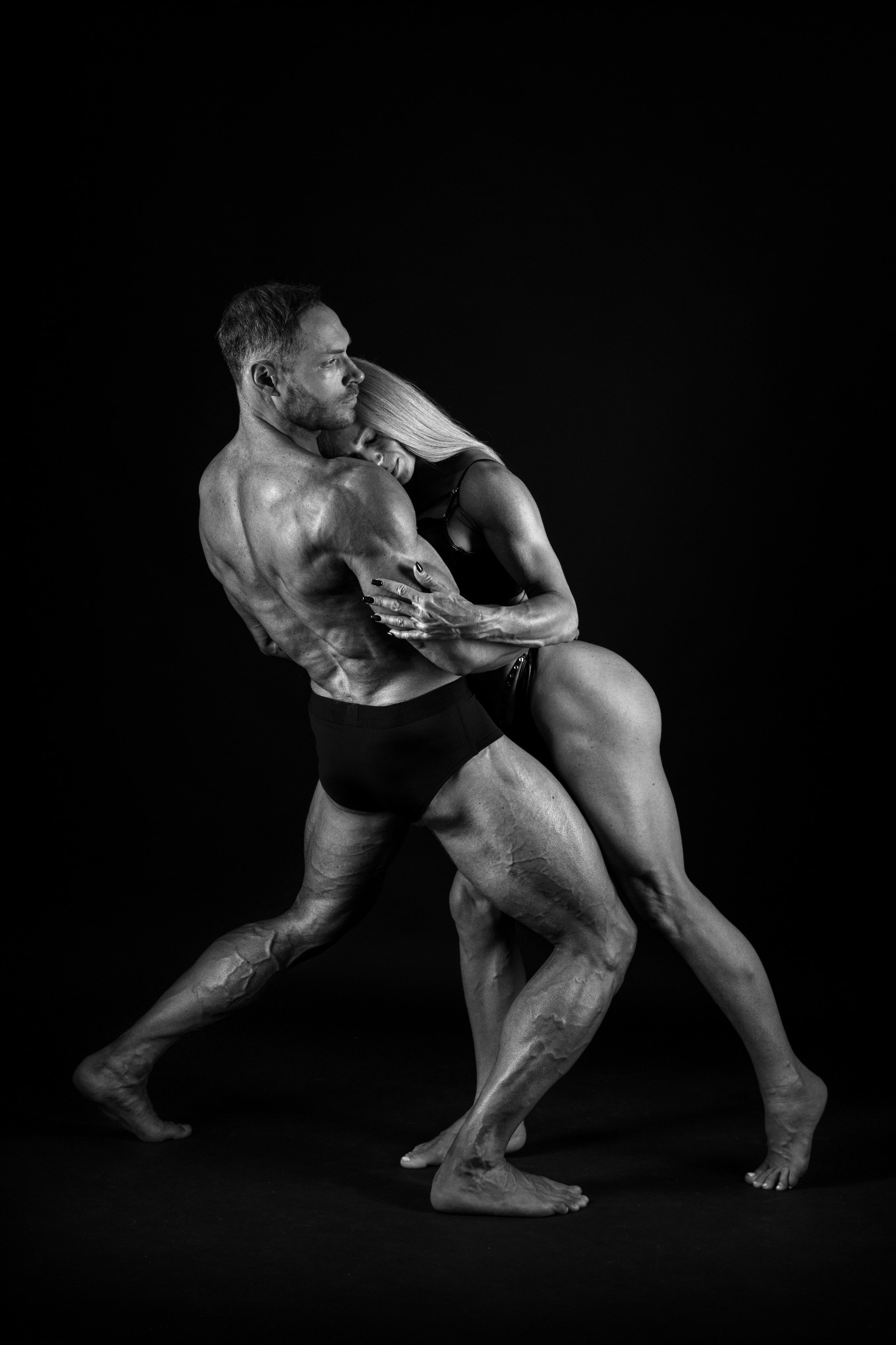 Czarno-białe zdjęcie muskularnego mężczyzny i kobiety w sportowej pozie na czarnym tle, ukazujące ich atletyczną sylwetkę i napięcie mięśni.