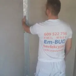 Pracownik w białej koszulce z logo firmy budowlanej przykłada długą poziomicę do świeżo nałożonego tynku na ścianie w narożniku pomieszczenia.