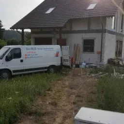 Dom w trakcie ocieplania elewacji, widoczny biały bus firmy budowlanej z otwartymi drzwiami, materiały budowlane obok budynku i świecące słońce.