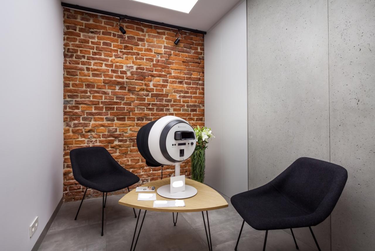 Wnętrze gabinetu z ceglaną ścianą, nowoczesnym urządzeniem diagnostycznym do skóry, stolikiem z wizytówkami i dwoma czarnymi krzesłami.