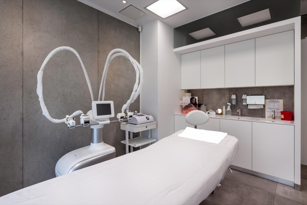 Wnętrze gabinetu medycyny estetycznej: białe łóżko zabiegowe z zagłówkiem, nowoczesny sprzęt do laseroterapii z osłonami na ramionach, białe szafki z umywalką i akcesoriami, szara ściana w tle.