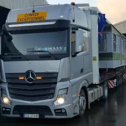 Srebrna ciężarówka Mercedes Actros z żółtą tablicą 'Convoi Exceptionnel' przewożąca mobilny dom na niskopodwoziowej platformie, widoczna tablica rejestracyjna RDE-27648.