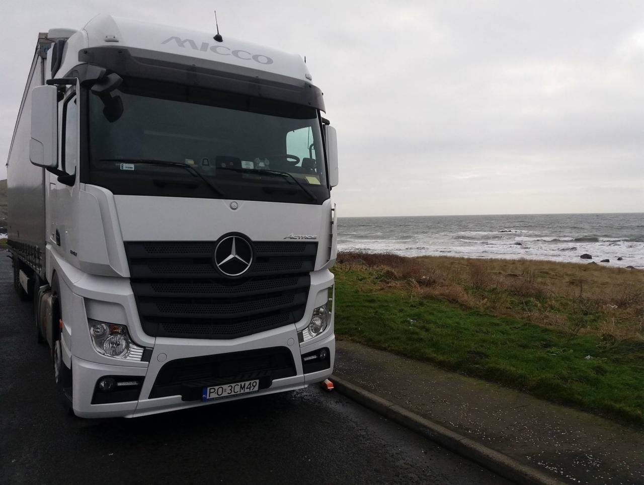 Biała ciężarówka marki Mercedes-Benz Actros z naczepą stoi na poboczu drogi, w tle widok na wzburzone morze i pochmurne niebo.