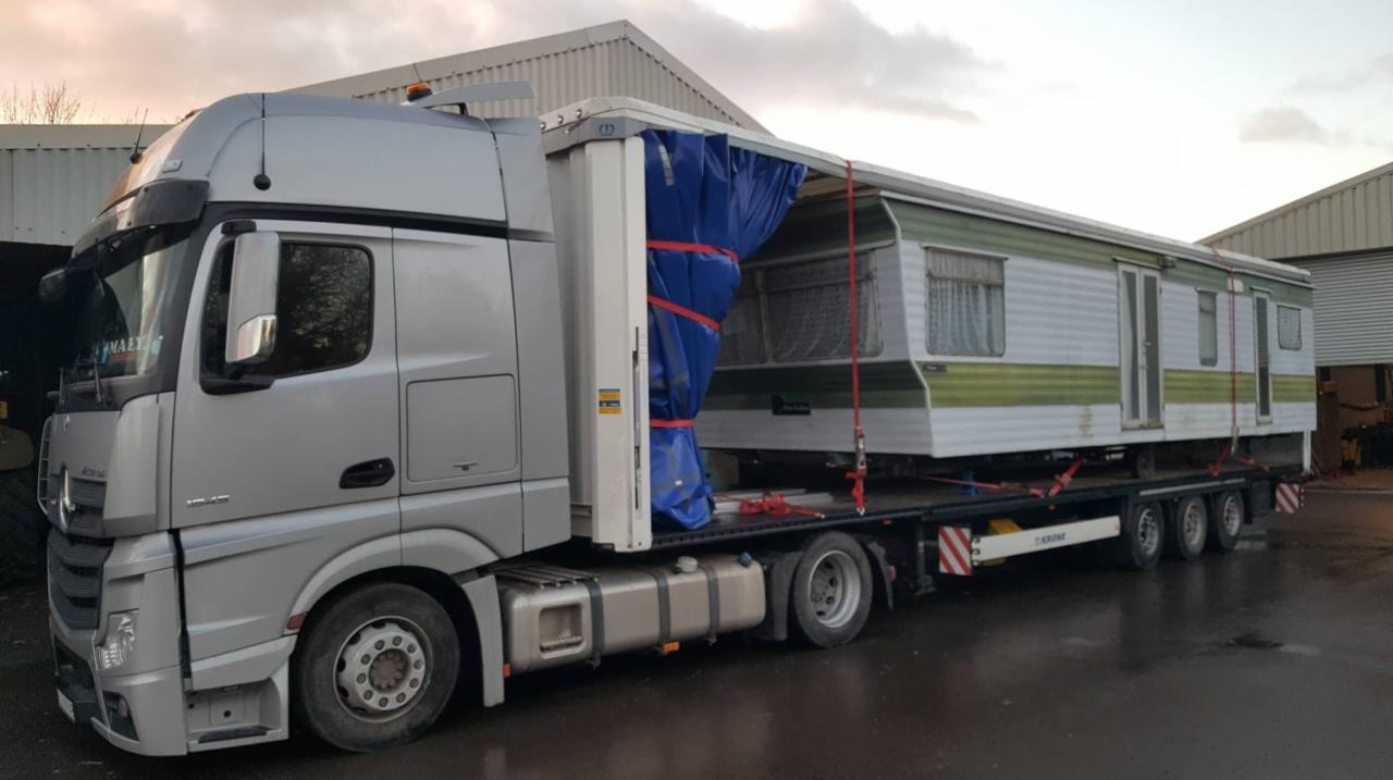 Srebrna ciężarówka Mercedes przewozi przyczepę kempingową z zielonymi akcentami, zabezpieczoną niebieską plandeką i czerwonymi pasami na niskopodwoziowej platformie.