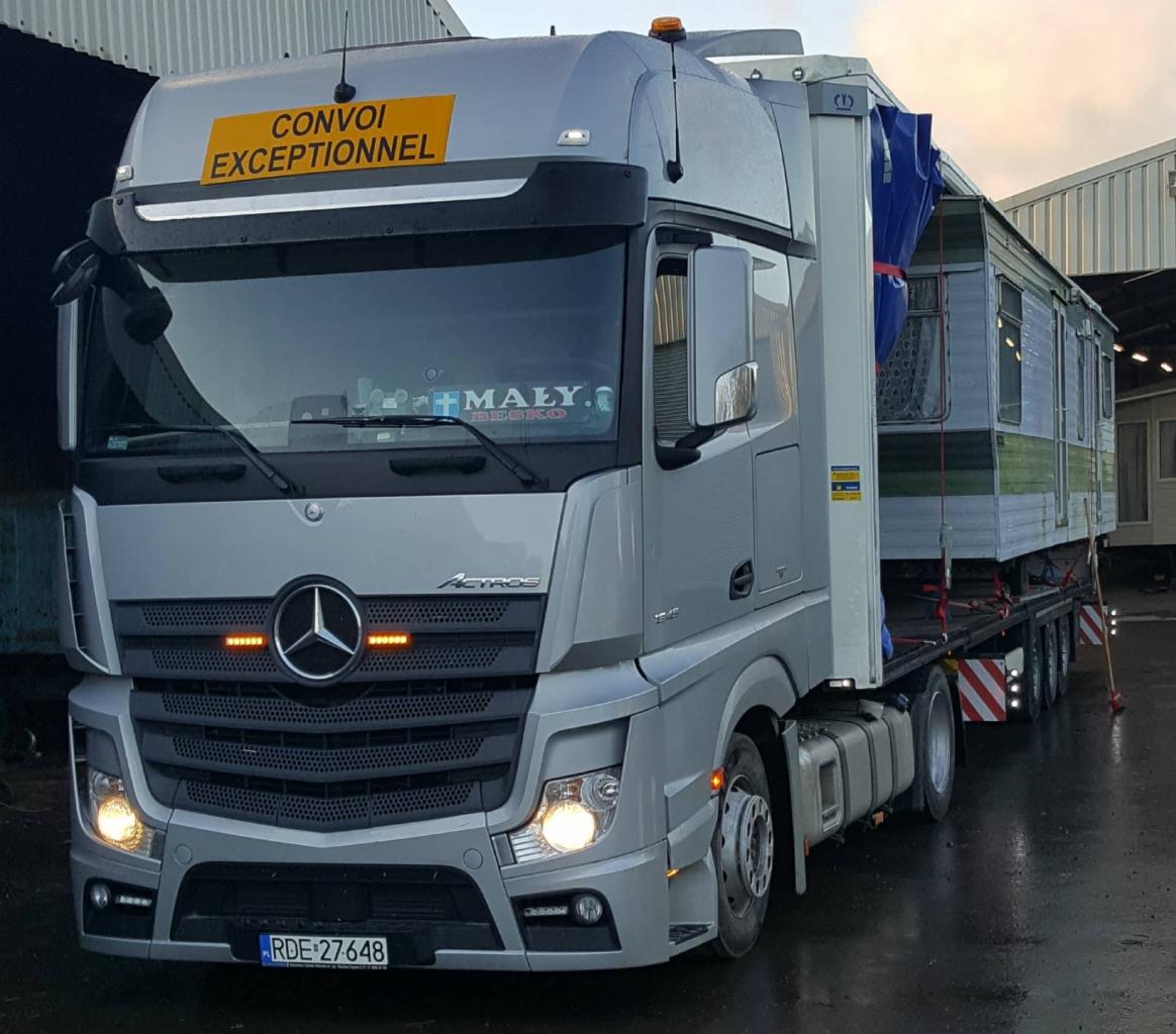 Srebrna ciężarówka Mercedes Actros z żółtą tablicą 'Convoi Exceptionnel' przewożąca mobilny dom na niskopodwoziowej platformie, widoczna tablica rejestracyjna RDE-27648.