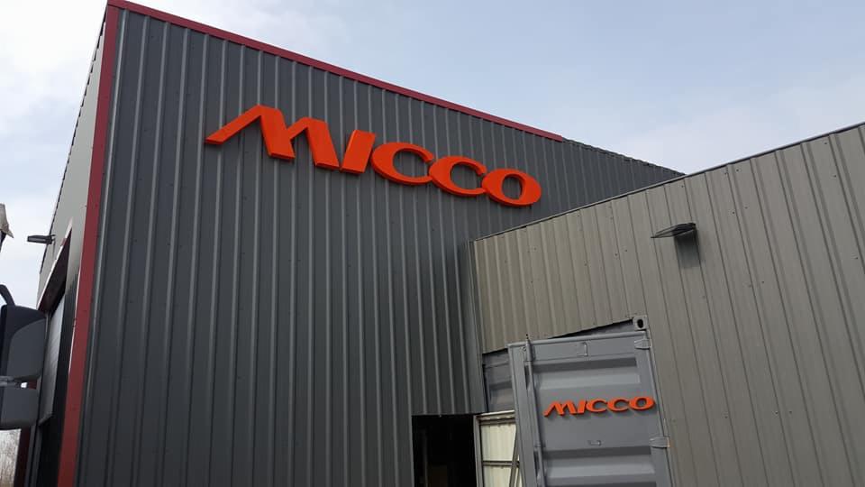 Budynek magazynowy z szarej blachy trapezowej z dużym pomarańczowym napisem Micco, z boku widoczny kontener z napisem Micco.