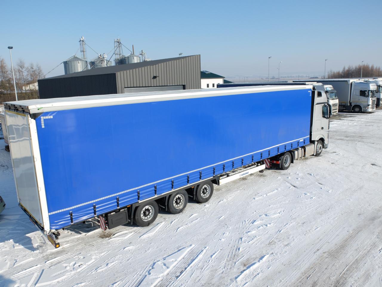 Niebieska naczepa ciężarówki na ośnieżonym placu, w tle silosy zbożowe i inne pojazdy.