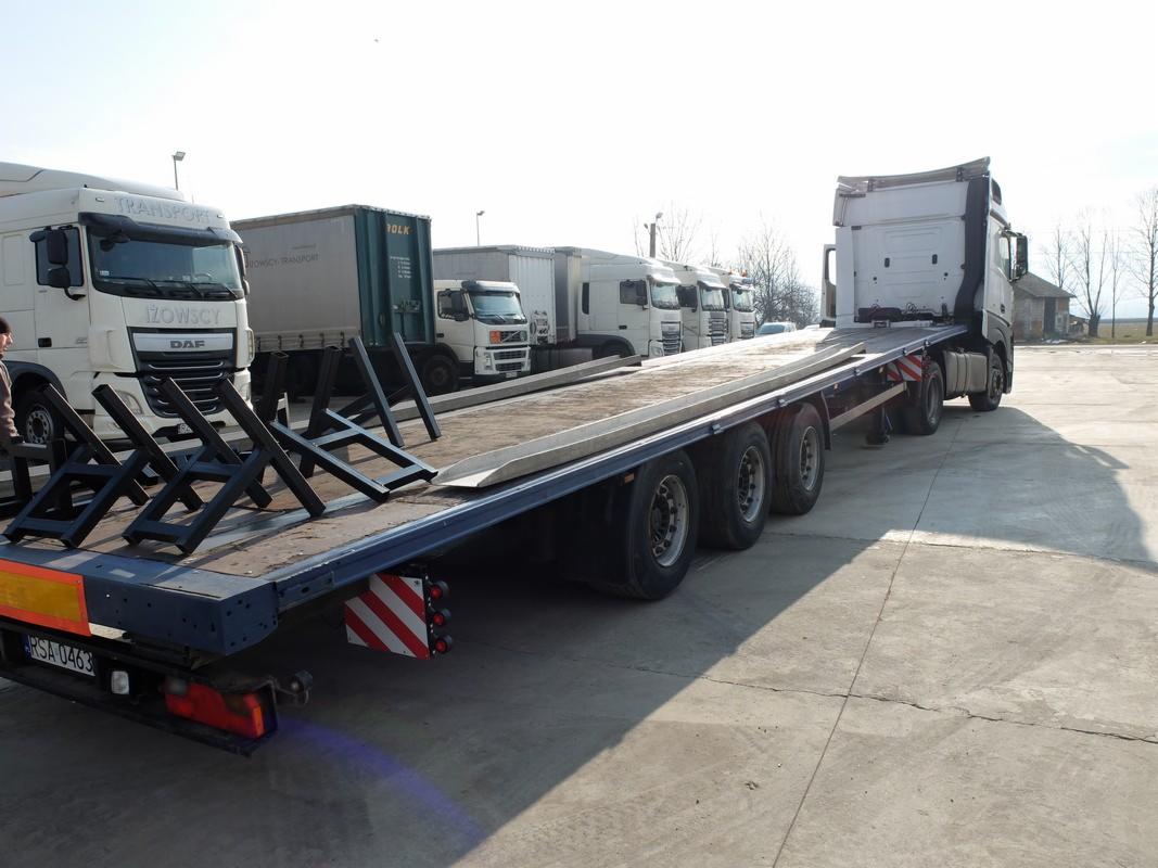 Ciężarówka niskopodwoziowa z rozłożonymi najazdami, zaparkowana na betonowym placu, w tle zaparkowane inne ciężarówki.