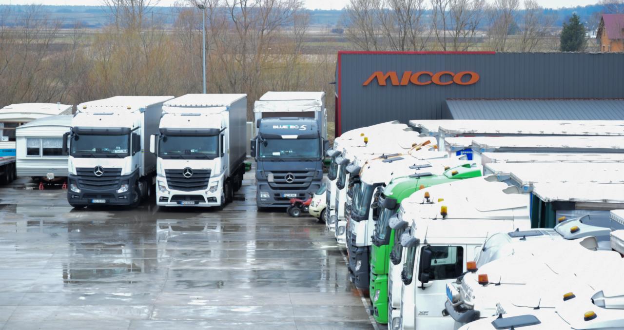 Flota samochodów ciężarowych Mercedes-Benz i innych marek zaparkowana na mokrym placu, z budynkiem firmy MICCO w tle.