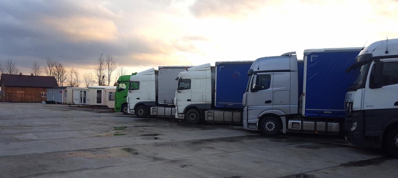 Rząd ciężarówek różnych marek i kolorów (zielony, biały, srebrny) z naczepami na placu, w tle baraki mieszkalne i budynek pod ciemnym, pochmurnym niebem.