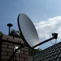 Antena satelitarna zamontowana na kominie z cegły na tle błękitnego nieba z chmurami, widoczny fragment dachu pokrytego czarną dachówką.