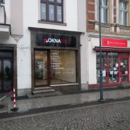 Fasada budynku firmy 'OKNAWRC' z witryną, na której widnieje oferta: okna, drzwi, rolety, żaluzje, parapety, bramy garażowe. Nad witryną szyld z nazwą firmy. Nad szyldem okno z brązową ramą. Ulica...