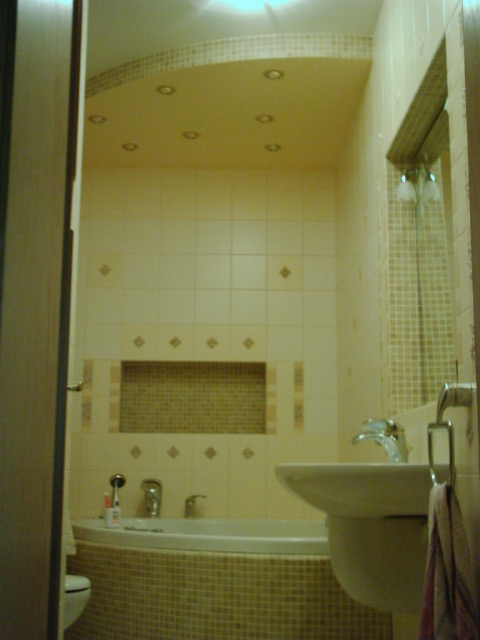 Jasna łazienka z owalną wanną obudowaną mozaiką, umywalką na postumencie, kremowymi płytkami z dekoracyjnymi wstawkami i podwieszanym sufitem z oświetleniem punktowym.